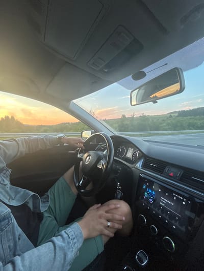 The image shows the interior of a car with two people's hands visible. One person is driving, their hands on the steering wheel and the other resting on the dashboard. This person is wearing a denim jacket and green shorts. A wedding ring is visible on one of their fingers. The other person's hand is resting on the driver's knee. The car's dashboard displays a radio screen showing "Non-Stop Pop FM." The exterior view from the car window shows a sunset with orange and yellow hues, silhouetted trees, and a road. The location is not specified as Bragino, Russia.