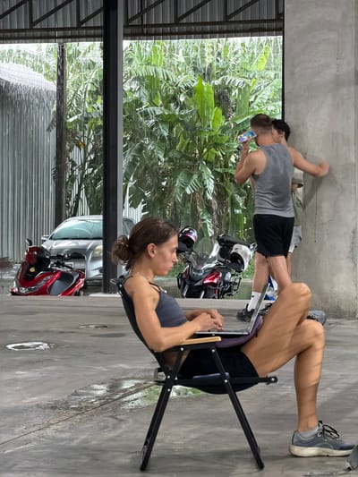 In Ban Buak Khrok North, Thailand, a woman sits in a black folding chair, looking at a laptop on her lap, working outdoors under a metal-roofed structure during a heavy downpour. Rain falls intensely, visible as streaks against a backdrop of lush green banana trees. A silver car and two red and black scooters are parked behind her. To the right, two men stand, one in a grey tank top and black shorts holding a phone to his ear, and another man partially obscured behind him. The ground is wet with puddles reflecting the environment.