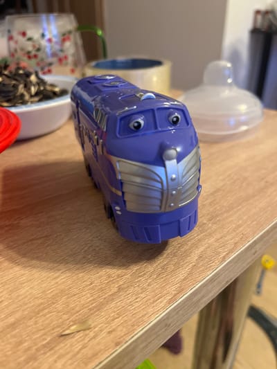 A purple toy train with large cartoon eyes and a silver grill on its front is placed on a light brown wooden surface. In the background, a white object resembling a baby bottle nipple is visible, along with a clear glass jar decorated with red cherries and a blue and white bowl filled with sunflower seeds. The scene appears to be indoors. No specific location in Pobiedziska, Poland, is identifiable from the image.