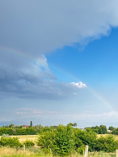 The image depicts a rural landscape in the Жібек жолы ауылдық округі, Kazakhstan, under a dramatic sky. A double rainbow is visible arcing across a sky with both dark, stormy clouds and patches of clear blue. Below the sky, a treeline of green foliage separates a yellow, harvested field from a distant range of hazy blue mountains. Several structures, including a building with a red roof and a tall, slender tree, are visible amongst the trees in the mid-ground. The foreground features dry grass and two wooden fence posts. The overall scene suggests a post-rain, transitional weather condition.