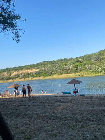 The image depicts a sunny day at a beach by a lake, possibly in Criuleni, Moldova. The foreground is dominated by a sandy beach with some sparse vegetation. In the mid-ground, people are engaged in various activities. Some are swimming or wading in the clear blue water, while others are relaxing on the sand. A beach umbrella provides shade for a small group of people.

The background features a lush green, tree-covered hillside rising from the edge of the lake. The water is calm with ripples suggesting a gentle breeze. The sky is a clear, bright blue, indicating fair weather. The overall scene conveys a sense of leisure and recreation.

Several people are visible, some in swimsuits. There are also some objects like kayaks or inflatable rafts in the water and on the shore. The time of day appears to be afternoon, given the angle of the sun and the bright lighting. There is no visible text in the image. The atmosphere seems relaxed and enjoyable, characteristic of a summer day at the beach.