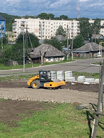 A yellow and black vibratory roller is operating on a dirt and gravel construction site in a suburban area. In the background, there are several multi-story apartment buildings, some older wooden houses with dilapidated roofs, trees, and a cellular tower. The scene is illuminated by natural daylight under a partly cloudy sky. The roller appears to be compacting the ground.