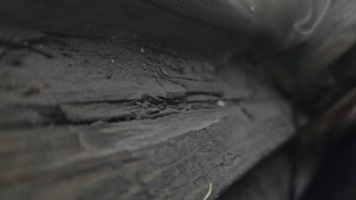This is a close-up, low-angle, macro photograph of weathered and cracked dark wood. The wood grain is prominent and appears rough and textured, with visible splinters and peeling layers. There is a thin layer of dust or dirt accumulated on the surface, especially in the crevices. The lighting is dim, creating deep shadows that emphasize the texture and age of the wood. The background is out of focus and consists of darker, undefined shapes, possibly other objects or the surrounding environment, with a hint of mesh-like material visible in the upper left corner. The scene is entirely devoid of any human presence or activity. The image does not contain any discernible elements that would allow for the localization of Kaka, Turkmenistan.