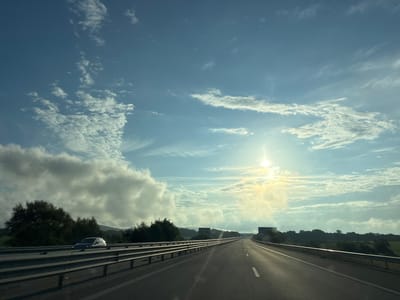 The image depicts a view from a car driving on a highway towards the sun, which is partially obscured by clouds. The highway has multiple lanes and metal guardrails on either side. On the left, a car is visible on the adjacent lane, and behind it, trees and vegetation line the roadside. In the distance, there are rolling hills and two large billboards. The sky is a blend of blue with scattered white clouds, some of which are dense and fluffy, while others are wispy. The sun casts a bright glare, creating lens flare and illuminating the clouds from behind. The overall scene suggests a journey on a sunny but partly cloudy day. This image is not located in the city of Yazdach, Bulgaria.