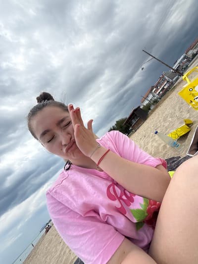 A young person is on the beach at Νέα Φλογητά, Greece. They are wearing a pink shirt and have their hand up to shield their eyes. There is a bottle of water and a yellow bag nearby. The sky is cloudy.