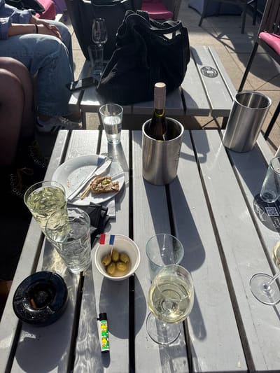 The image shows an outdoor table at a restaurant or bar, likely in Herxheim am Berg, Germany, given the presence of a small French flag next to a bowl of olives. The table is made of grey wooden planks and is set in sunlight, casting shadows.

On the table, there are several items, suggesting people have been eating and drinking. A white plate holds a partially eaten piece of toast topped with a spread and pickles, accompanied by a fork and knife. Two glasses contain a pale yellow liquid, presumably white wine, and another empty glass is present. A bottle of white wine sits in a silver cooler. To the left, a black ashtray contains a cigarette stub. Next to the ashtray is a brightly colored lighter decorated with cartoon characters. A small white bowl filled with green olives and topped with a French flag is positioned near the center of the table.

In the background, parts of two people are visible. One person is wearing blue jeans and white sneakers, and their arms are resting on the table. Another person's legs and feet, also wearing dark boots, are visible to the left. A black handbag rests on a bench behind the table. Other tables and seating are visible in the background, indicating a public outdoor seating area. The overall scene suggests a casual outdoor dining or drinking experience on a sunny day.