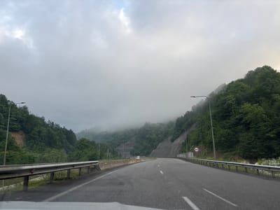 The image shows a view from a vehicle traveling on a two-lane highway that curves to the right. The highway is bordered by guardrails on both sides and concrete barriers in the median. The surrounding environment is mountainous and heavily forested with lush green trees. There is a noticeable fog or low-lying clouds obscuring the upper portions of the mountains. On the right side of the road, a circular speed limit sign indicating "80" is visible. A white van can be seen in the distance on the highway. The sky is overcast with diffused light. The road surface appears to be asphalt. The scene is likely captured in a rural mountainous region of Georgia, given the presence of the speed limit sign and the distinctive landscape.