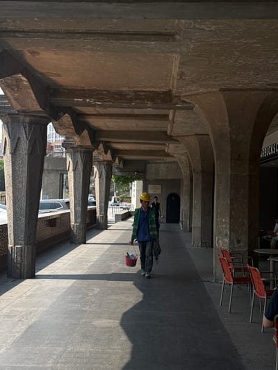 A man wearing a yellow hard hat and blue clothing walks through a stone arcade in Hongkou District, China. There are red chairs and a sign for "Starbucks" visible at the end of the arcade. The arcade columns are made of stone with detailed carvings.