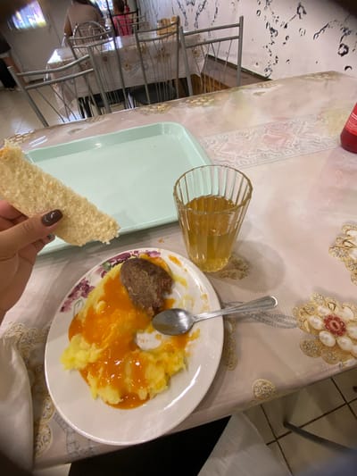 The image shows a close-up of a meal on a table covered with a patterned tablecloth. A white plate contains mashed potatoes topped with an orange sauce and a piece of meat. A spoon rests on the plate. To the right, a glass of amber-colored liquid is visible. In the foreground, a hand is holding a piece of white bread. In the background, a light blue tray is on the table, and other people are seated at tables in what appears to be a canteen or dining hall. The environment suggests a casual dining setting in Akyar, Russia.