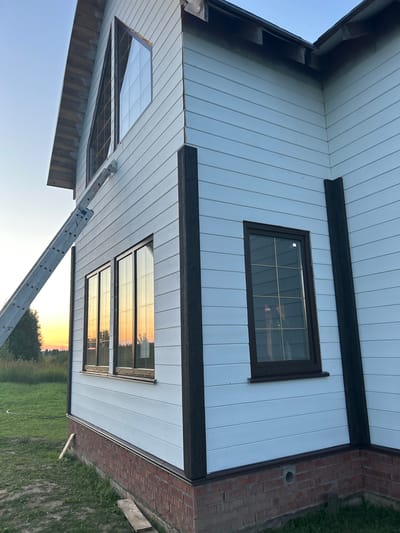 The image depicts the exterior of a two-story house under construction in Abushkovo, Russia. The house features light blue horizontal siding, dark brown trim around windows and vertical corner supports, and a red brick foundation. Several windows are visible, including a large triangular window on the upper level and multiple rectangular windows on the lower level. A silver ladder is propped against the side of the house, extending towards the upper window. The surrounding environment consists of a green grassy lawn with some trees and vegetation in the background. The sky is clear and shows the warm hues of a sunset. There are no people visible in the image.