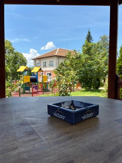 The image shows a dark blue ashtray with several cigarette butts inside, sitting on a wooden table. The ashtray has the word "Victory" written on its sides in white lettering with a gold outline. In the background, out of focus, is a children's playground with colorful slides and structures, a house with a red roof, and lush green trees under a bright blue sky with scattered clouds. The scene is viewed from under a wooden overhang, suggesting a covered outdoor seating area, possibly a patio or porch. The specific location in Anton, Bulgaria, cannot be determined from the image alone.
