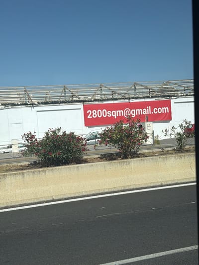 The image shows a bright, sunny day in Can Negre, Spain. A large, red banner is displayed on the side of a white building, with white text that reads "2800sqm@gmail.com". The building appears to be an industrial or commercial structure, with an exposed metal framework on its roof. In front of the building, there is a line of green bushes with pink flowers, and a few white vehicles are parked or driving in the background. A concrete barrier runs along the foreground, separating the building area from a dark asphalt road with white lane markings. The sky is a clear blue. There are no people clearly visible in the image.