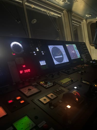 The image shows a ship's bridge at night. Various control panels and displays are visible, including radar screens, gauges, and digital readouts. A compass repeater and engine order telegraph are prominent. The room is dimly lit with illuminated buttons and screens. Windows are on the left side of the image.