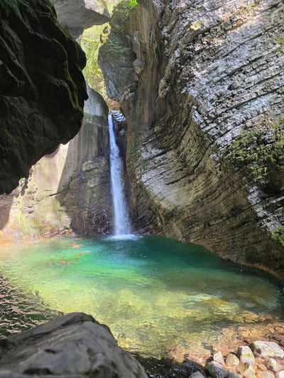 This image depicts the Kozjak waterfall and pool within a cave-like setting near Kobarid, Slovenia. The waterfall plunges into a pool of clear turquoise water, contrasting with the surrounding grey and tan rock formations. The walls of the cave are layered and textured, showing signs of erosion and moisture. Some parts of the rocks are covered with moss. A large rock mass dominates the lower left corner of the frame, its surface heavily textured with moss. Sunlight filters into the cave, illuminating sections of the water and the rock faces. The composition is oriented on its side with the waterfall on the left and the pool on the right.
