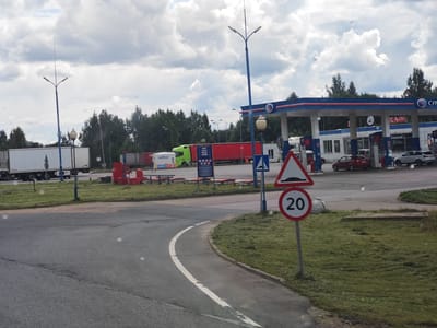 The image depicts a roadside scene near the city of Kresttsy, Russia. In the foreground, a paved road curves to the left, bordered by a grassy verge. A white line delineates the edge of the road. On the right, a speed limit sign indicates "20" within a red circle, and a triangular warning sign with a speed bump symbol is also visible.

In the background, a gas station is prominent, featuring a blue and red canopy over fuel pumps. Signs with Russian lettering, including "КАФЕ," are attached to the station building. Several vehicles are present, including a red car, a white SUV, and a white truck with a trailer. Further to the left, a green truck and another white truck with a trailer are parked. A lamppost with multiple lights stands near the center of the scene. Trees line the background, and the sky is filled with white, puffy clouds. The overall impression is a typical roadside service area.