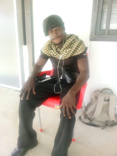 A dark-skinned man wearing a black headwrap, a black t-shirt, and black pants sits on a red plastic chair outdoors. He has a plaid scarf around his neck. White earbuds are in his ears and the cord drapes down his chest. A small black bag is worn across his chest. He wears black shoes and sits with his legs crossed. To his right, on the ground, is a light gray backpack. The background consists of a white wall with vertical paneling, and to the right, a window with horizontal blinds. The floor is a light-colored concrete. The image appears to be taken in Indo, Nigeria.