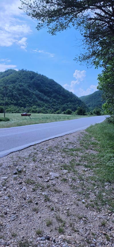 This image captures a rural landscape in Echinos, Greece, under a clear blue sky with scattered white cumulus clouds. A light grey asphalt road extends through the mid-ground, subtly curving to the right in the distance. A solid white line delineates the left edge of the road, which is flanked by gravel shoulders on both sides.
To the left of the road, a large green grassy field stretches towards a series of densely forested hills that form the dominant background. Within this field, a single wooden utility pole stands upright near the road. Further into the field, a red pickup truck is visible, oriented away from the viewer, with its rear section and truck bed discernible amidst the green grass.
The right side of the frame is characterized by an overhanging tree, its dark branches and green foliage partially obscuring the upper right portion of the sky. The immediate foreground on the right consists of a gravel and dirt shoulder, sparsely covered with small rocks, patches of green grass, and small white flowers. There are no individuals or animals present, and no actions are observed within the scene.