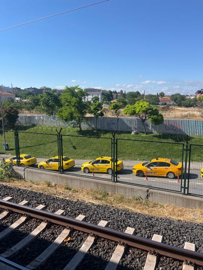In Zeytinburnu, Turkey, a line of five yellow taxis is parked adjacent to a metal fence topped with barbed wire. The taxis are positioned on a paved area bordered by a concrete retaining wall. Behind the fence, a grassy slope rises towards a backdrop of urban buildings and lush green trees under a clear blue sky. The foreground features a section of railroad tracks with dark gray ballast and concrete sleepers, one of which has a small orange object resting on it. The scene appears to be captured from a slightly elevated perspective, looking down at the parked vehicles and the railroad.