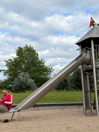 The image depicts a playground slide structure with a metal slide. A man wearing a red hooded jacket and tan pants is sitting at the bottom of the slide, looking at his phone. A small black object, possibly a speaker, is on the sand next to him. The playground is covered in sand, and a grassy area with trees is visible in the background. The sky is cloudy. The location is the playground in the Slipe in Boxtel, Netherlands.