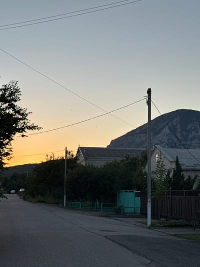The image depicts an outdoor scene, likely in a rural or semi-rural setting, with a road leading towards a residential area. A power pole with wires runs diagonally across the frame, suggesting the presence of electricity infrastructure. In the background, a large mountain looms, its texture and form visible against the sky. Adjacent to the road, there is a building with a light-colored facade and a dark-colored roof, partially obscured by trees. A green metal fence or gate is also visible near the building. The sky exhibits a gradient of colors, transitioning from a pale blue at the top to a warm yellow or orange near the horizon, indicating that it is either sunrise or sunset. The overall mood is tranquil, with a hint of natural beauty provided by the mountain and the sky. There are no people visible in the frame. The image appears to be taken during the golden hour.