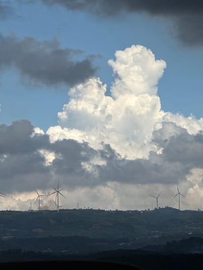 The image depicts a scenic view of rolling hills with several wind turbines positioned on the crest of the landscape. The sky is dramatic, filled with a mix of dark, heavy clouds and bright, puffy cumulus clouds, suggesting changing weather conditions. In the foreground, the rolling hills are covered in dense, dark green vegetation, with some hints of structures and possibly roads visible on their slopes. The wind turbines, with their tall white towers and rotating blades, are silhouetted against the sky and the clouds. The overall scene captures a sense of vastness and the power of nature, with the wind turbines representing a human intervention in this natural environment. The specific location is identified as Đa Lộc, Vietnam.