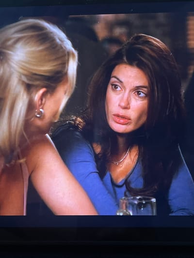 This media file shows two women, one with dark hair and the other with blonde hair, in an indoor setting. The woman with dark hair is closer to the camera and is looking upwards with a surprised or concerned expression. She has dark, wavy hair and is wearing a blue shirt and a silver necklace. The blonde-haired woman is in the foreground, with her back to the camera, and her head is tilted upwards. She is wearing a silver hoop earring. The background is blurred but appears to be a dimly lit interior, possibly a bar or restaurant, with hints of wooden elements and possibly other patrons. The scene suggests an intimate or intense moment between the two women. There is a glass on a table in the background, with what appears to be a reddish liquid in it. The overall lighting is dim, which could indicate an evening setting. No text is visible in the image.