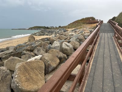 The image shows a wooden boardwalk with dark grey grated decking, curving upwards along a rocky shoreline. The boardwalk has brown wooden railings. To the left of the boardwalk is a beach composed of sand and large, rough rocks. Beyond the rocks, the sea is visible with waves breaking on the shore. In the distance, across the water, there is a pier with some structures and buildings on a grassy hill. The sky is overcast and grey. The scene is located in Courtown, Ireland, along the coast.