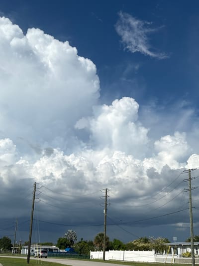 A bright, overcast day is depicted in North Port, Florida, with a dramatic sky dominating the frame. Large, fluffy white cumulus clouds fill the upper portion of the image, set against a deep blue sky. Below these, a layer of darker, ominous storm clouds stretches across the horizon, suggesting an approaching weather system.
In the foreground, a paved road runs horizontally, bordered by a grassy median. Three wooden utility poles, strung with numerous power lines, stand tall along the side of the road. The poles are weathered and show signs of age.
On the left side of the road, a silver SUV is parked. Beyond the road and a white picket fence, a low-rise building with a light-colored roof is visible, along with scattered palm trees and other greenery. The overall impression is a suburban or semi-rural landscape under a dynamic sky.