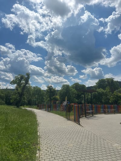 The image depicts a park or playground area with a paved walkway in the foreground. To the left of the walkway is a grassy area with scattered wildflowers. The walkway curves to the right, leading towards a playground enclosed by a colorful, multi-colored fence. Playground equipment, including slides and climbing structures, is visible beyond the fence. Several wooden benches are also present within the fenced area. In the background, lush green trees form a dense backdrop, with a hazy hillside visible further in the distance. The sky above is a vibrant blue, dotted with large, fluffy white and grey clouds, suggesting a bright, partly cloudy day. The overall scene conveys a sense of an outdoor recreational space within a natural environment. The location is the city of Donchovci, Bulgaria.