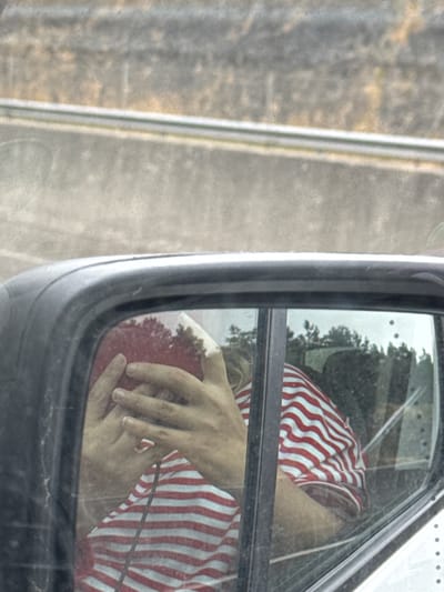 The image shows a reflection in a car's side mirror of a person wearing a red and white striped shirt. The person is covering their face with their hands, and a red object, possibly a hat, is visible. The background of the reflection shows trees and foliage. The image is taken from inside the car, looking out through the side mirror. The setting is a road with a concrete barrier. The specific location in Dunbell, Ireland, cannot be determined from the provided image.