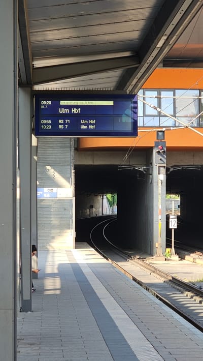 The image shows a train station platform in Neu-Ulm, Germany. A digital display board lists train departures. The top entry shows "09:20 RS 7" with a delay of approximately 5 minutes and the destination "Ulm Hbf". Below that, at "09:55 RS 71 Ulm Hbf" and "10:20 RS 7 Ulm Hbf". A person is seated on a bench on the platform, looking at their phone. The platform is made of concrete tiles with textured strips. A train tunnel is visible in the background, with tracks curving into the darkness. Overhead electrical wires and a traffic signal for trains are also present. The sun is casting long shadows, indicating it is likely late afternoon or early morning. The building in the background is orange.
