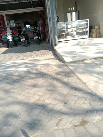 The image captures a street scene in Kemamang, Indonesia, featuring a bakery and a display case. In the foreground, a paved area with interlocking bricks shows dappled sunlight and shadows. To the left, a bakery with a red accordion-style door is partially open, revealing the interior. A man in a black shirt stands near the counter inside. Three motorcycles are parked in front of the bakery, two of them appearing to be the subject of recent activity. A person wearing a grey hooded jacket sits on one of the motorcycles. In the middle ground, a glass display case filled with various items is positioned outside a building to the right. The case has multiple shelves, and the items on display are not clearly identifiable but appear to be packaged goods. Behind the display case, boxes are stacked, some with labels. The background shows a building with a grey wall and a doorway. The overall scene suggests a commercial area with outdoor elements.