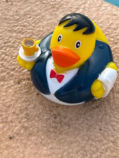 A rubber duck toy, designed to resemble a duck wearing a suit and holding a cup of coffee, rests on a textured, sandy surface. The duck is primarily yellow with a black hairstyle, a red bow tie, and a navy blue suit. Its wings are depicted as yellow with white cuffs. The sandy surface appears to be outdoors, possibly near a body of water given the presence of what looks like a blue surface in the upper right corner of the image. The lighting suggests it is daytime. The exact location in Sant Vicenç de Montalt, Spain, cannot be determined from the image alone.