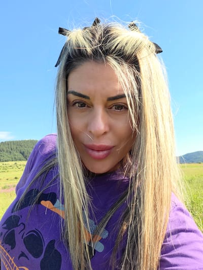 A woman with blonde hair, wearing a purple t-shirt with a skull graphic, is posing for a selfie outdoors. She is positioned in the foreground, with a clear blue sky and a green, hilly landscape in the background. The landscape features fields and a forest. The image appears to be taken on a sunny day.