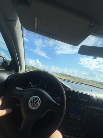 The image is a first-person perspective from inside a Volkswagen vehicle, driving on a paved road on a sunny day. The steering wheel, with the Volkswagen logo, is prominently in the foreground, and the dashboard with the radio and instrument cluster is visible. The view through the windshield shows a rural landscape with fields, trees, and hills under a bright blue sky with scattered white clouds. A distant car and a triangular warning sign are visible on the road ahead. The lighting suggests daytime. The image was taken in the vicinity of Svetlina, Bulgaria.