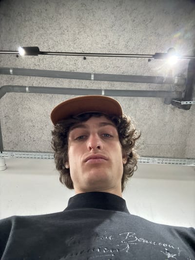The image displays a male individual photographed from a low-angle perspective, looking directly upwards into the camera. He has brown, curly hair and wears a brown baseball cap with the brim facing forward. He is dressed in a black high-neck long-sleeved top featuring light-colored embroidery or print on the chest. His expression is neutral. The background consists of a textured, light grey concrete ceiling. Exposed utility infrastructure is visible, including multiple dark grey pipes secured by metal clamps and brackets, running horizontally across the upper portion of the frame. A horizontal metal track or conduit is also present, equipped with at least two bright light fixtures, one of which on the right exhibits significant lens flare. The environment indicates an indoor, utilitarian setting, consistent with a parking garage, basement, or service area. The specific location within Joinville-le-Pont, France, is not identifiable from the visual content; however, the scene details suggest an urban infrastructure environment.