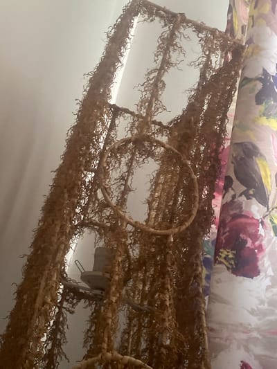 A close-up, slightly blurry, vertical shot shows a hanging macrame plant holder, possibly made of dried moss or twine, with a circular hoop in the middle. The plant holder hangs against a plain, off-white wall, with a floral patterned curtain visible on the right side. The lighting suggests an indoor environment, possibly a room. There are no people visible in the image.