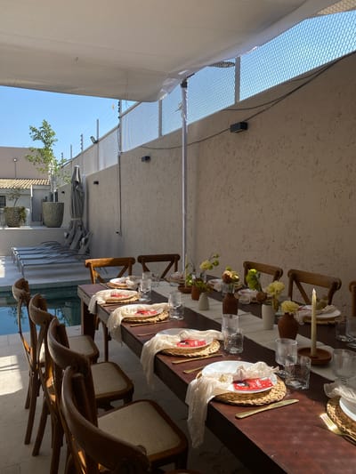 The image shows an outdoor dining area, likely a restaurant patio, in what could be Santa Cruz Tehuixpango, Mexico, judging from architectural styles. A long, dark wood table is set for a formal meal. The table features place settings with woven chargers, white plates, glassware, cutlery, and decorative name cards marked "Charly Grad." Several small floral arrangements in vases are spaced along the center of the table. The table is surrounded by wooden chairs with upholstered seats and backs. The area is covered by a translucent roof structure, possibly a combination of metal mesh and canvas shading. In the background, hints of buildings, landscaping, and patio umbrellas suggest the location is part of a commercial area. The scene appears to be prepared for a celebration or special event.