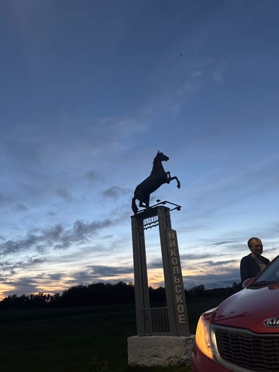 A silhouette of a horse statue stands on a tall pedestal against a dusky sky in the city of Nikolskoye, Russia. The statue depicts a rearing horse. Below the horse, on the pedestal, is a sign with Russian text that reads "НИКОЛЬСКОЕ" vertically. To the right of the pedestal, a man with glasses is partially visible, leaning against a red Kia car. The car's headlights are illuminated, casting a warm glow. The background shows a dark silhouette of trees and a field under a sky with streaks of clouds and remnants of sunset colors. The overall scene is set outdoors at dusk.
