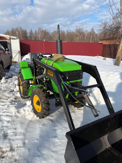 A bright green mini-tractor with a black front-end loader and bucket attachment is parked on snow-covered ground in Уром, Russia, under a clear blue sky with scattered white clouds. The tractor features bright green body panels, black fenders, and yellow wheel hubs on black, deeply-treaded tires. Tire chains are visible on the rear wheels. A prominent black exhaust pipe rises vertically from the engine compartment. The front-end loader frame is black, with the Cyrillic word "БАТЫР" (BATYR) embossed in yellow on its side. A light-colored fabric strap is secured over the tractor's engine cover. To the immediate left of the tractor, a red car, possibly an SUV or crossover, is partially visible, parked near a light-colored building with a white door. In the background, a tall, reddish-brown corrugated metal fence extends horizontally. Behind the fence, a dense line of bare deciduous trees stands under the clear sky. To the far right, a section of a bare tree trunk is visible. The ground is covered unevenly with white snow, showing areas of compacted or partially melted snow. No people are present in the scene.