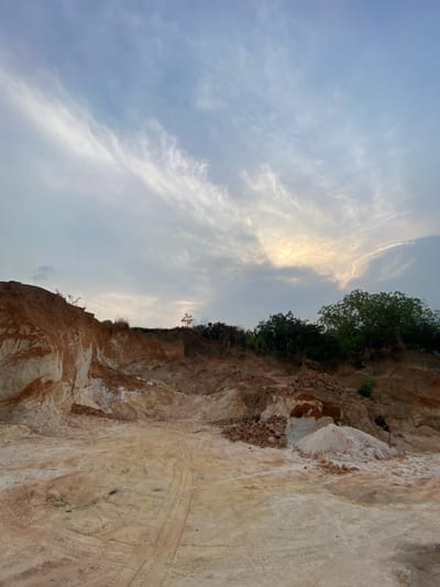 The image depicts an active or recently active open-pit excavation site in Asaga Amangwu, Nigeria. The ground surface features exposed layers of reddish-brown soil and lighter, off-white sedimentary material, with distinct vehicle tire tracks visible in the foreground. The excavation is bordered by steep, stratified earthen banks that display similar soil color variations. Sparse green vegetation and trees are present along the upper edges of these banks, particularly on the right side. In the lower right background, a solitary individual, dressed in dark clothing, is observed in a bent posture, seemingly engaged in manual labor with a tool near a pile of excavated material. The sky is predominantly overcast with mixed cloud formations, and a bright, diffuse light source, indicative of the sun, is visible through the clouds in the upper right quadrant.