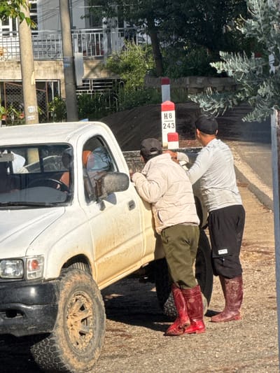 Two men stand next to a white pickup truck on a dusty road in Tuý Sơn, Vietnam. The man on the left, wearing a light-colored jacket, olive-green pants, and red boots, leans his head against the truck. The man on the right, in a light grey long-sleeved shirt and black pants, has his right hand on the truck and his left hand on the shoulder of the other man. A red and white marker post with "H8 243" is visible behind them. The truck appears dirty, with mud on its exterior and tires. In the background, a building with balconies and greenery can be seen.