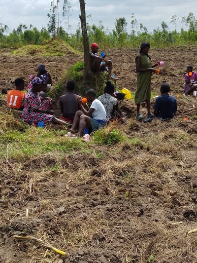 A group of people gathers near a field with a tree. Some sit on the ground, while others stand. They appear to be taking a break, holding cups or food. A woman stands in the middle, perhaps distributing drinks. The field shows signs of recent tilling, with exposed soil and remnants of harvested crops. Trees and vegetation surround the field, indicating a rural setting.