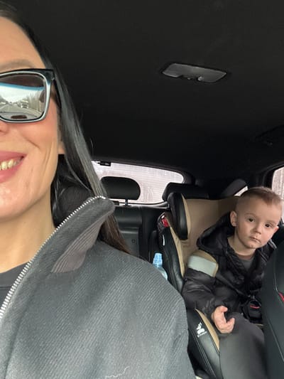 The scene depicts the interior of a vehicle located in Kabanov Mys, Russia. In the immediate foreground, a portion of an adult, identified as female, is visible on the left side of the frame. She wears dark-framed sunglasses, reflecting a view of a road and trees, and has a slight smile. Her long dark hair is visible, and she is wearing a dark, zippered jacket. To the right and slightly behind, a young male child, estimated to be between three and six years old, is seated in a beige and black child booster seat in the rear passenger compartment. The child is dressed in a dark puffer jacket and looks directly towards the viewer with a neutral expression. A plastic water bottle with a blue cap is positioned to the left of the child's booster seat. The vehicle's interior features a dark headliner and dark upholstery on the seats.