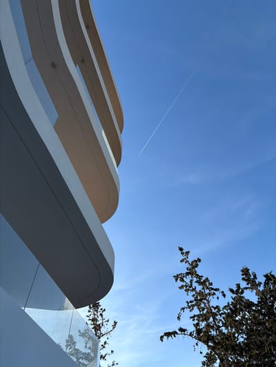 The image presents a low-angle view of a contemporary architectural structure, likely located in the Municipal Unit of Elliniko, Greece. The building features a series of curvilinear, cantilevered balconies or awnings, constructed from a smooth, likely concrete or composite material. The surfaces are light-colored, possibly white or a light grey, and reflect the ambient light. Clear glass panels are visible between the curved elements. The sky is a clear blue with light cirrus clouds and a visible contrail from a jet plane. A tree with dark foliage is positioned in the upper right corner of the frame, partially obscuring the sky. The photograph appears to be taken during daylight hours under sunny conditions.