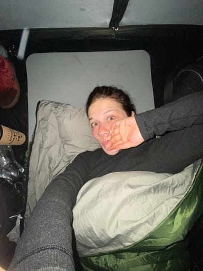 A young woman is lying in a sleeping bag inside a tent. She is wearing a dark gray long-sleeved shirt and dark gray pants. Her hair is dark brown and pulled back. She is looking at the camera with her mouth slightly open and her right hand covering her mouth. There is a white pillow under her head. To the left of the sleeping bag is a gray mat, and to the left of that is a red shoe and a brown cylindrical object with writing on it. The tent appears to be set up in an outdoor location. The image was taken in Абзаново, Russia.