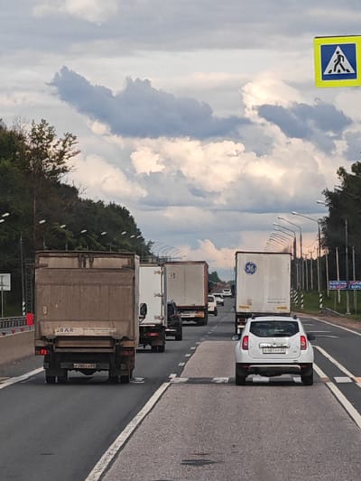 The image shows a busy highway in Kolesnye Gorki, Russia, with several trucks and cars on the road. The sky is cloudy, with dramatic cloud formations. In the foreground, a white SUV is driving in traffic. Behind it, a brown truck is visible. Further down the road, more trucks and cars are present. A pedestrian crossing sign is visible on the right side of the road. The environment appears to be a semi-urban or suburban area, with trees lining the sides of the highway.