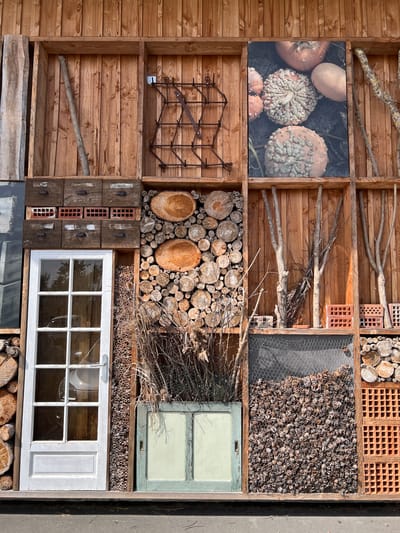 This media file presents a detailed, multi-compartment structure, likely an insect hotel or an artistic installation, crafted primarily from natural and recycled materials. The scene is depicted as a rustic, vertical grid of various textures and forms, mounted on or constructed from wood.
The structure is composed of wooden shelves and partitions creating numerous distinct sections. In the upper left, compartments are filled with arrangements of tree branches and twigs. Below these, a framed picture displays a close-up of several warty, textured pumpkins in shades of orange and cream. Further down, a metal grid or trellis is mounted, and the bottom-most left compartment contains a single, thick branch resting on a shelf.
Moving to the central and right sections, a variety of materials are meticulously arranged. One area features neatly stacked cut logs and wood slices of varying diameters. Adjacent to these are compartments filled with terracotta bricks, characterized by their perforated, grid-like structure. Bundles of dried twigs and reeds are also present, some forming dense clusters. A notable section on the right is enclosed by chicken wire and densely packed with numerous small pine cones.
Further down, two old window frames are incorporated: one with a pale blue-green frame in the upper right-middle, and a larger white-framed window at the bottom right. Below the blue window, a section is filled with many thin, hollow tubes, likely reeds or bamboo, typical components of an insect hotel. In the bottom-middle, dark, weathered wooden blocks are interspersed with smaller sections of perforated terracotta bricks.
The overall lighting is bright and natural, suggesting a clear day, highlighting the textures of the wood, bark, and other elements. No discernible text is visible on any of the objects. The setting implies an outdoor location, consistent with the provided context of Loire-Authion, France, emphasizing sustainability and natural aesthetics.