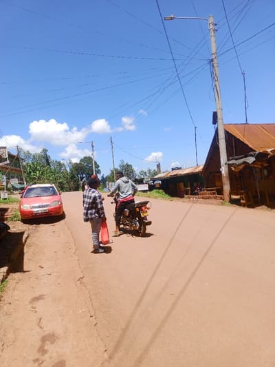 The image depicts a daytime street scene in Kahuro, Kenya, under a clear blue sky with scattered white cumulus clouds. A reddish-brown dirt road, likely unpaved, occupies the central and foreground areas.
In the foreground-left, an individual wearing a plaid shirt and light-colored pants stands, facing away from the camera, holding a bright orange/red bag. Next to them, slightly to the right, another individual is seated on a dark-colored motorcycle, also facing away, wearing a grey hooded sweatshirt and red shorts/pants. The motorcycle's rear license plate, "KMA...", is partially visible.
On the left side of the road, a red Toyota Corolla sedan (license plate KBK 168P) is parked on the dirt shoulder, facing the viewer. Further left, green foliage and a building with a green facade and red roof are partially visible.
To the right of the road, multiple utility poles with numerous intersecting overhead wires are prominent. One pole features a street lamp. Several single-story buildings line the right side of the street; these structures have rusted corrugated metal roofs and simple facades, indicative of small shops or residences. Individuals are visible sitting outside one of these buildings in the background.
The scene conveys typical activity along a main street in a rural or semi-urban Kenyan settlement.