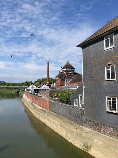 This is an outdoor shot of Harvey's Brewery in Lewes, United Kingdom. The brewery complex includes buildings with dark gray slate roofs and white-framed windows, along with brick sections. A notable chimney rises from the center of the complex. There's a body of water, possibly a river, reflecting the buildings and sky. In the sky, there are scattered clouds and two birds are visible. The brewery's signboard, showing its name and establishment date (1790), is prominently displayed. The scene is viewed from an elevated position, possibly a bridge or a high vantage point.